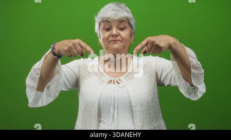 Frau, Seniorengröße mit grauem Haar, zeigt beide Zeigefinger beim Lächeln nach unten, trägt weiße Bluse und Armbänder im Studio; verspielte Invita Stockfoto