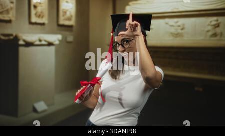 Frau hält Diplomrolle mit rotem Band und zeigt Zeigefinger auf Kappe, während sie in einem Museumsgebäude lächelt; stolz. Stockfoto