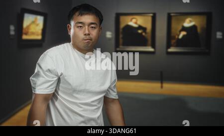 Junger chinesischer Mann in weißem T-Shirt steht still und starrt auf gerahmte Gemälde an der Wand im schwach beleuchteten Galeriebau; Nachsicht. Stockfoto