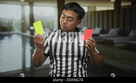 Junger chinesischer Schiedsrichter hält gelbe und rote Karte neben dem Innenpool im Gebäude. Stockfoto