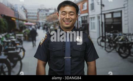 Mann in Polizeiuniform steht lächelnd mit Badge und Radio an der Schulter auf der Straße neben geparkten Fahrrädern; Stolz-Dienst. Stockfoto
