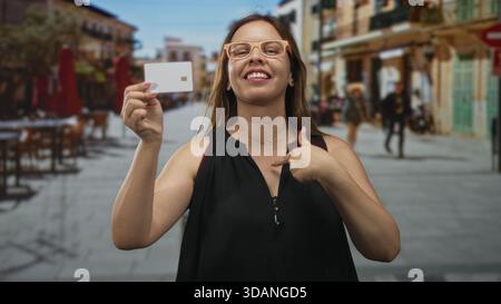 Frau, die eine Kreditkarte hält und mit dem Finger auf die Brust zeigt, während sie auf der Straße lächelt und eine Brille trägt; Selbstbewusstsein. Stockfoto