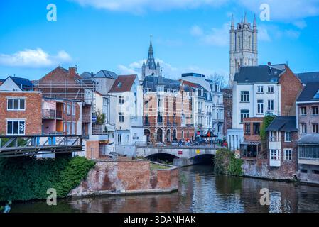 Ein Kanal in Gent ist gesäumt von Ziegelbauten und weißen Gebäuden, einer Brücke mit Menschen, die überqueren, und hohen Kirchtürmen, die sich im Hintergrund unter einem bri erheben Stockfoto