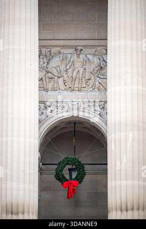 Washington Planning The Battle Of Trenton Relief William Jefferson Clinton Federal Building Washington DC | WASHINGTON — Ein Weihnachtskranz hängt unter dem Relief „Washington Planning the Battle of Trenton“, eingerahmt zwischen geschwungenen dorischen Säulen im Andrew W. Mellon Auditorium in Washington, D.C. Der Bildhauer Edmond Amateis hat die Tafel aus Indiana-Kalkstein im Jahr 1936 gemeißelt, um George Washington darzustellen, wie er seinen Angriff auf Trenton plant, flankiert von den Generälen Nathanael Greene und John Sullivan, deren Gesichter als versteckte Porträts des Architekten des Gebäudes und eines Mitbildhauers gestaltet sind. Es gehört zu dem Federal Triang Stockfoto