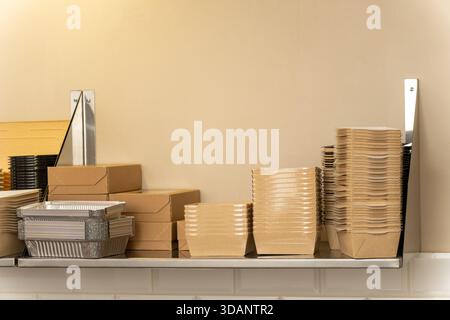 Verschiedene Einweg-Lebensmittelverpackungsartikel, einschließlich Pappbecher, Kartons und Behälter, die in einem Metallregal in einer kommerziellen Umgebung ausgestellt werden Stockfoto