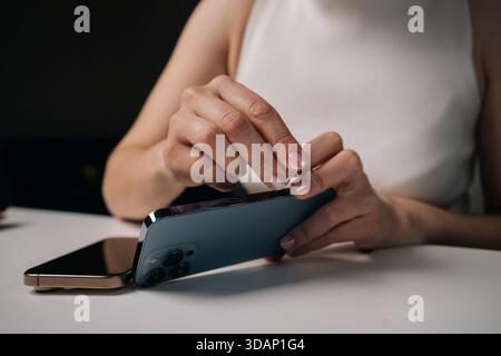 Frau, die an einem weißen Tisch sitzt und das sim-Auswerferwerkzeug verwendet, um die sim-Kartenablage des Mobiltelefons zu öffnen, und die sich mit der Verwaltung der Gerätekonnektivität beschäftigt. Stockfoto
