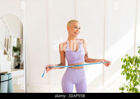 Schlankheitskonzept. Junge Frau, die Taille mit Klebeband misst, ihre Körperparameter überprüft, Sportbekleidung trägt Stockfoto