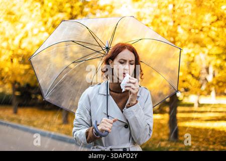 Frau im Regenmantel mit Regenschirm isst Snack im Herbst im Park mit gelben Blättern Stockfoto