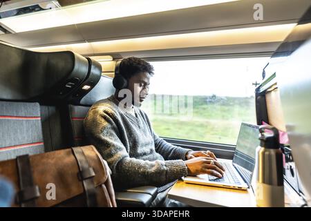 Junger Mann mit Kopfhörern, der sich auf seinen Laptop konzentriert, unterwegs arbeitet oder auf Geschäftsreisen unterwegs ist, während er mit dem Zug unterwegs ist und sich konzentriert und professionell entwickelt Stockfoto