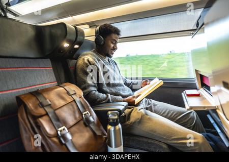 Junger schwarzer Mann, der in einem modernen Zug reist, Kopfhörer trägt und ein Buch liest, und mit seinem Rucksack eine komfortable und produktive Reise genießt Stockfoto