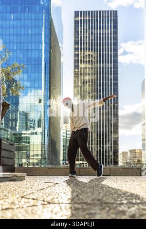 Junger Mann mit roter Mütze, der in einer dynamischen Pose tanzt, während er in einer urbanen Stadtlandschaft tanzt, hinterleuchtet vom Sonnenlicht zwischen hoch aufragenden Glashochhäusern auf einem s Stockfoto