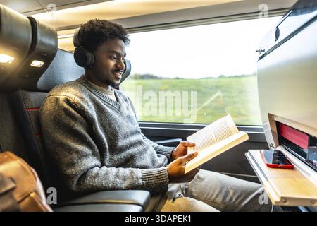 Junger Mann, der Kopfhörer trägt und ein Buch liest, während er bequem mit dem Hochgeschwindigkeitszug reist, während er seine persönliche Zeit und Unterhaltung genießt Stockfoto