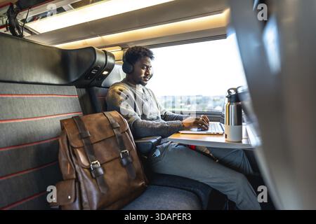 Junger Mann, der Kopfhörer trägt und mit dem Zug an einem Laptop arbeitet, der sich auf Fernarbeit und den Lebensstil digitaler Nomaden konzentriert und sich auf einen komfortablen Aufenthalt freut Stockfoto