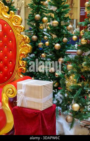 Eine festliche Weihnachtspräsentation im Innenbereich mit einem markanten rot-goldenen Weihnachtssessel, einem gestreiften Geschenk und wunderschön dekorierten Weihnachtsbäumen. Stockfoto