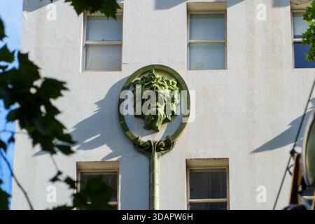 Aus nächster Nähe eine kunstvoll verzierte Skulptur mit grünem Löwenkopf, die ein strukturiertes weißes Gebäude ziert, eingerahmt von Fenstern und Baumblättern. Stockfoto