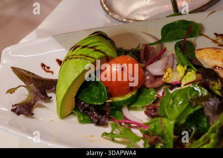 Bei einer Nahaufnahme wird ein frischer und lebendiger Salat aufgenommen, der fachmännisch mit Avocado, einer ganzen Tomate und verschiedenen Grünsorten zubereitet wird. Stockfoto