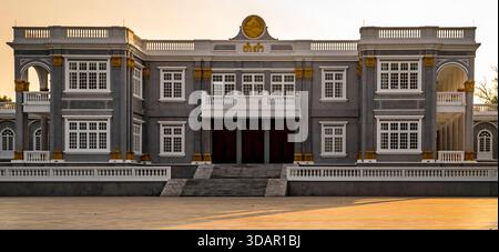 Ein großes, symmetrisches graues Gebäude mit weißen Fenstern und goldenen Akzenten, mit einem zentralen Eingang und einer breiten Treppe unter klarem Himmel. Stockfoto