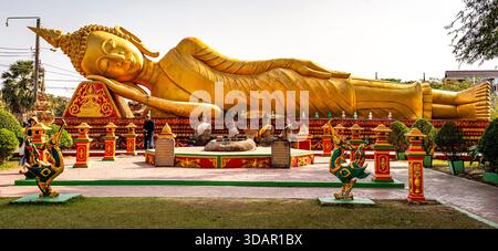 Goldene liegende Buddha-Statue in einem Tempelgarten mit kunstvoller Dekoration und Grün unter klarem Himmel. Stockfoto