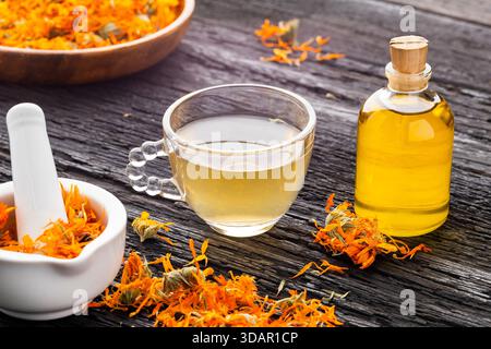 Calendula Tee und ätherisches Öl auf einem rustikalen Tisch, begleitet von getrockneten Blütenblättern und natürlichen Utensilien - Calendula officinalis Stockfoto