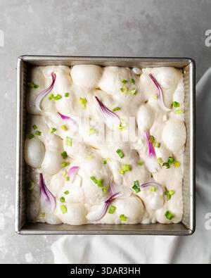 Draufsicht von Zwiebeln- und Schalottenfokaccia-Teigscheiben in einer quadratischen Backform, Draufsicht von luftiger, sprudelnder Zwiebelfokaccia-Teigprobung Stockfoto