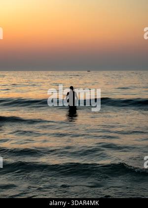 Silhouette einer Person, die bei Sonnenuntergang in der Nähe von Chania, Kreta, Griechenland, im Meer steht, mit ruhigem Wasser und warmem Abendlicht. Stockfoto