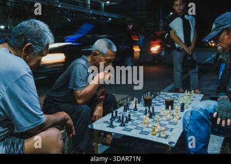 Balikpapan, Indonesien - 1. November 2025. Eine Gruppe von Männern, die sich intensiv auf ein ernsthaftes Schachspiel konzentrieren, das nachts an einer Straßenecke gespielt wird. Stockfoto