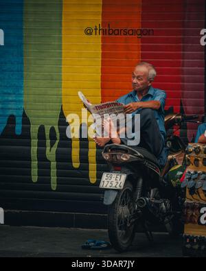 Ho-Chi-Minh-Stadt, Vietnam November 2025 ein älterer Mann sitzt auf einem Motorrad und liest eine vietnamesische Zeitung vor einem bunt bemalten Rolltor auf einem Stockfoto