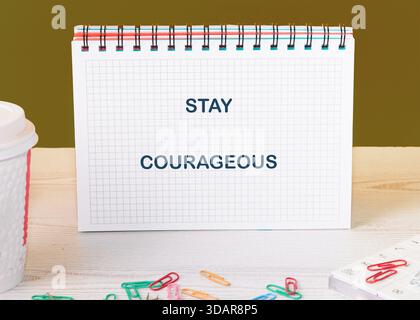 Ein Notizblock zeigt die Worte STAY COURAGEOUS auf einem weißen Holztisch. Bunte Büroklammern sind in der Nähe zusammen mit einer Kaffeetasse verstreut, wodurch ein vib entsteht Stockfoto