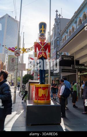 Melbourne, VIC, Australien. Dezember 2025. - Eine große Nussknacker-Soldat-Kunstinstallation auf einer roten Trommel ziert einen lebhaften Bürgersteig in der Bourke Street in mir Stockfoto