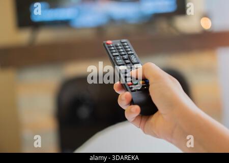 Hand mit tv-Fernbedienung zum Einschalten des Fernsehers zu Hause. Rückansicht einer Frau auf dem Sofa, die den Fernsehsender wechselt. TV-Fernbedienungskonzept. Selektiver Fokus Stockfoto