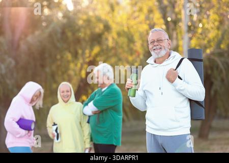 Sportlicher Seniorenmann mit Fitnessmatte und Wasserflasche gegen Gruppe im Park Stockfoto