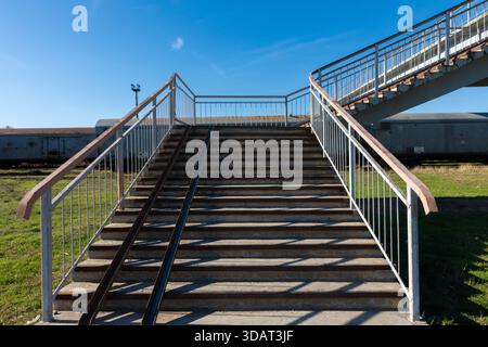 Betontreppe mit Metallschienen, die zu einer Fußgängerbrücke über Eisenbahngleise unter klarem blauem Himmel führt. Stockfoto