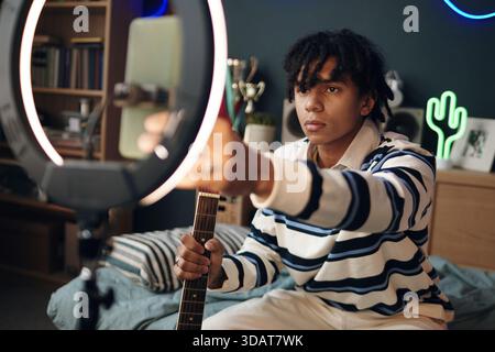 Porträt des schwarzen Teenagers, der auf dem Bett sitzt und akustische Gitarre hält und das Ringlicht anpasst, während er in die Kamera blickt, Schlafzimmer Hintergrund mit Bücherregalen und Dekor sichtbar Stockfoto