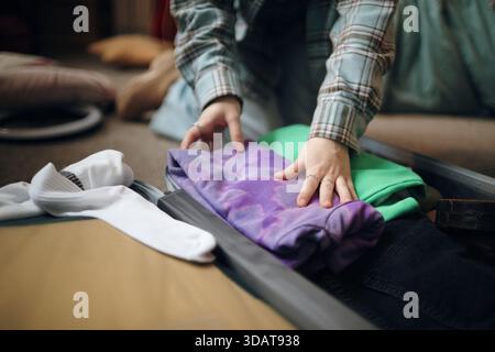 Kaukasische junge Erwachsene Frau packt Koffer, faltet Kleidung und organisiert Gegenstände für Reisen, Hände sichtbar arrangieren Kleidung, bereitet sich auf die Reise im Innenbereich vor Stockfoto