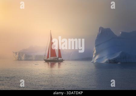 Der Blick auf das Segelboot mit roten Segeln navigiert durch eiskaltes Wasser neben kolossalen Eisbergen unter einem trüben, goldenen Himmel, Grönland. Stockfoto