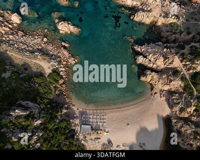 Blick aus der Vogelperspektive auf den goldenen Sand, der auf das türkisfarbene Wasser trifft, eingerahmt von zerklüfteten Felsen und üppigem Grün in Sardinien, Italien. Stockfoto