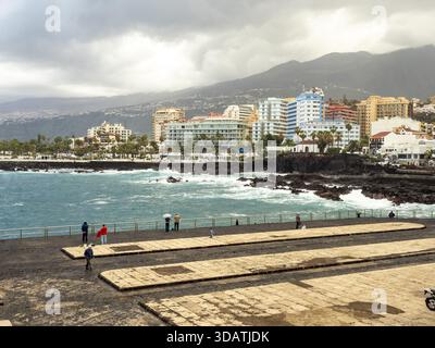Blick auf den dunklen Pier, der sich in das türkisfarbene Meer erstreckt, im Kontrast zu den hellen Hotels vor der bergigen Kulisse, Puerto de la Cruz, Tener Stockfoto
