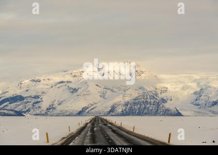 Der Blick auf eine lange, dunkle Straße führt durch eine riesige, schneebedeckte Ebene zu einer majestätischen, schneebedeckten Bergkette unter einem weichen, bewölkten Himmel, Island. Stockfoto
