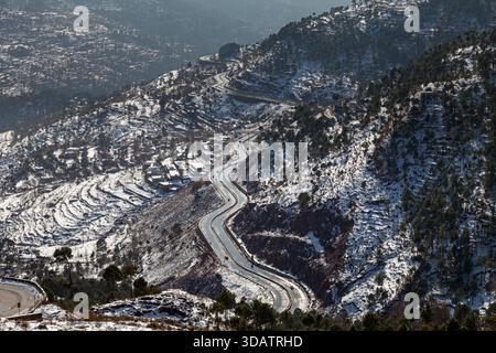 Blick auf eine gewundene Straße schlängelt sich durch schneebedeckte Hügel, ein steifes Asphaltband, das sich von der unberührten weißen Landschaft abhebt, Murree, Punjab, Pakista Stockfoto