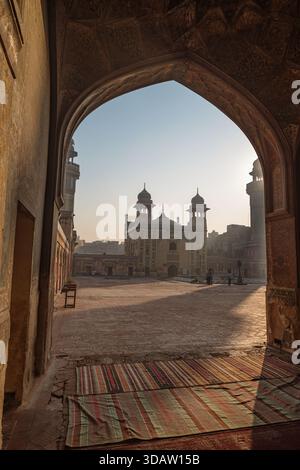 Blick auf die majestätische Badshahi-Moschee, eingerahmt von einem bogenförmigen Eingang, der Schatten auf den Innenhof mit warmem Sonnenlicht wirft, Lahore, Punjab, Pakistan. Stockfoto