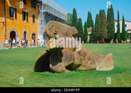 Pisa, Italien - 26. Juni 2025: Eine Skulptur des gefallenen Engels liegt über dem Gras der Piazza dei Miracoli, deren verwitterte Form vor Touristen steht Stockfoto