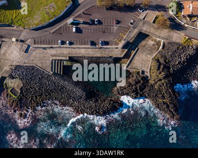 Aus der Vogelperspektive dunkle vulkanische Felsen, die das türkisfarbene Wasser in der Nähe der Betonstrukturen von Capelas, Azoren, Portugal umschließen. Stockfoto