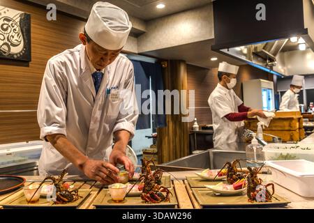 In der Küche eines Ryokan-Restaurants in Toba, Präfektur Mie, organisiert ein Koch mehrere Ise-EBI (japanischer Hummer, Panulirus japonicus) als Sashimi auf lackierten Tabletts. Im Hintergrund arbeiten andere Köche. Das Ise-EBI gilt als Delikatesse der Mie-Region und ist ein zentrales Merkmal des Kaiseki-Menüs in Ryokans auf der Ise-Halbinsel. Toba, Mie, Japan Stockfoto