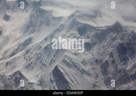Aus der Vogelperspektive sehen Sie majestätische, schneebedeckte Berge, die durch eine Wolkendecke durchdringen, ihre zerklüfteten Gipfel und frostigen Hänge, die eine krasse, winterliche Pfanne bilden Stockfoto