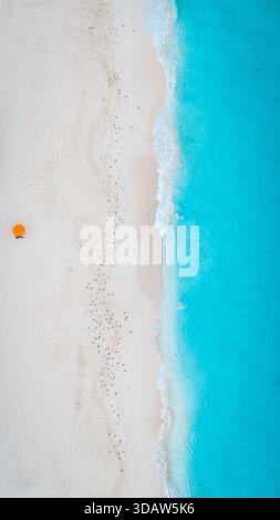 Aus der Vogelperspektive auf türkisfarbene Wellen, die sanft den unberührten weißen Sand küssen, wo ein einsamer orangener Regenschirm herausragt, Zakynthos, Griechenland. Stockfoto