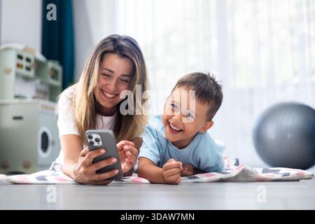Lächelnde Mutter und Sohn liegen auf dem Boden, kleben über ein Smartphone. Der fröhliche Moment fängt Lachen und Konnektivität in einem gemütlichen Wohnzimmer setti ein Stockfoto