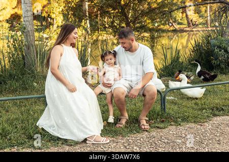 Eine multinationale Familie genießt einen ruhigen Tag im Park. Die schwangere Mutter schließt sich ihrem Mann und ihrer Tochter an, während sie in der Nähe von Enten am Teich, shari, sitzen Stockfoto