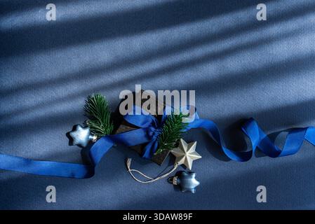 Eine wunderschön gewickelte schwarze Geschenkbox, die mit einem leuchtend blauen Band gebunden ist und von grünen Kiefernzweigen und metallischen Sternornamenten akzentuiert wird, sorgt für ein festliches H Stockfoto