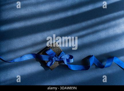 Eine wunderschön verpackte Geschenkbox mit einem auffälligen blauen Band sitzt auf einer weichen, strukturierten blauen Oberfläche. Die elegante Präsentation erinnert an festliche Ho Stockfoto