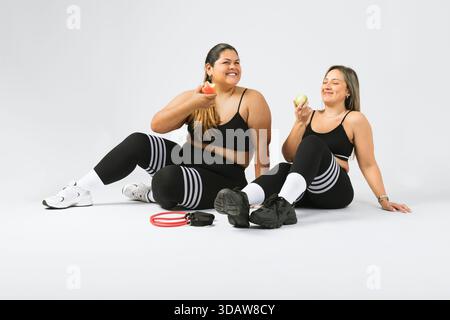Zwei Frauen sitzen in Aktivkleidung auf dem Boden, lächeln und genießen Äpfel. Sie verkörpern eine körperpositive Botschaft, die Wohlbefinden und Vertrauen in A unterstreicht Stockfoto